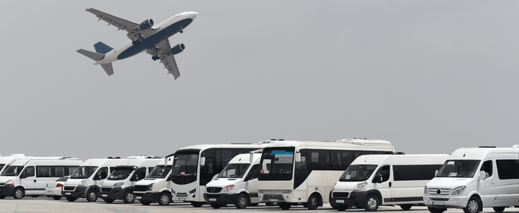 Minibuses and vans lined up on an tarmac while a plane takes off overhead